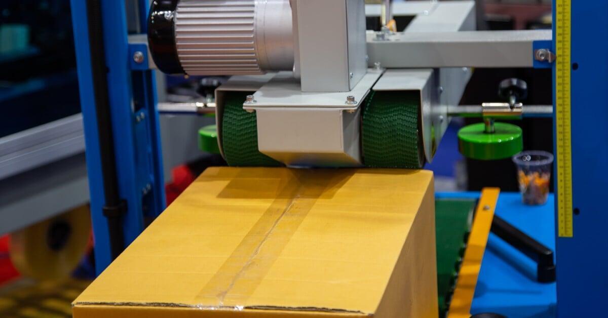 An automatic case sealer machine applies a strip of clear packing tape to a large yellow cardboard box.