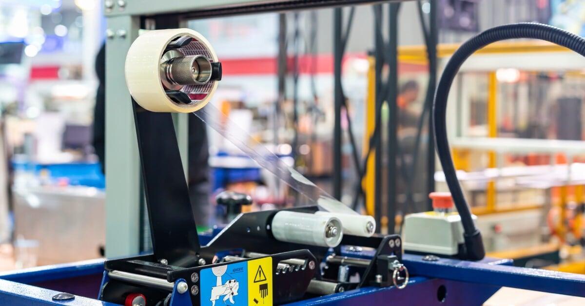 A length of clear plastic tape stretches from the roll to a small roller in a commercial case sealing machine.