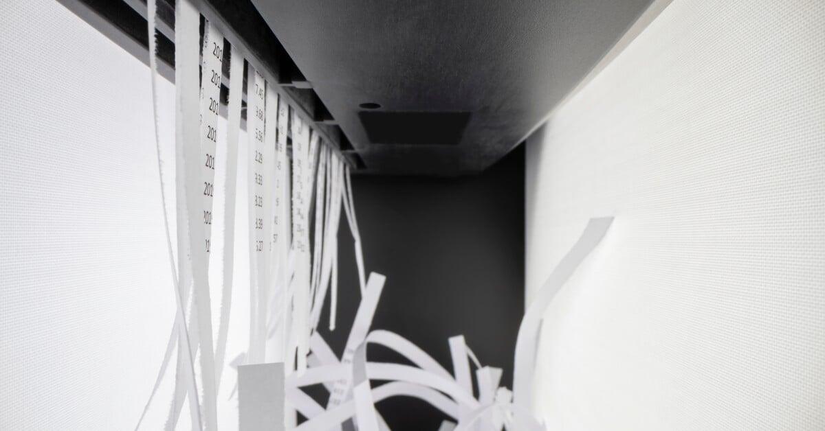 Thin strips of paper emerge from an industrial paper shredded machine. The paper features long lines of numbers.