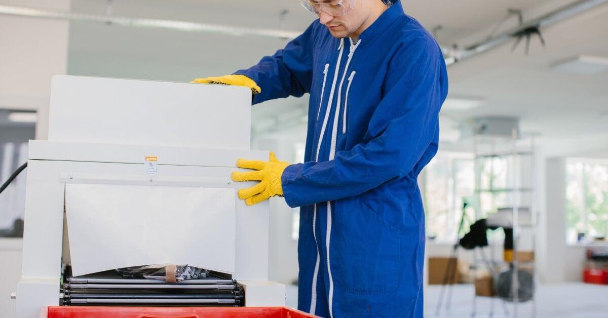 A warehouse worker wears a blue jumpsuit and safety gloves while operating an industrial paper shredder.