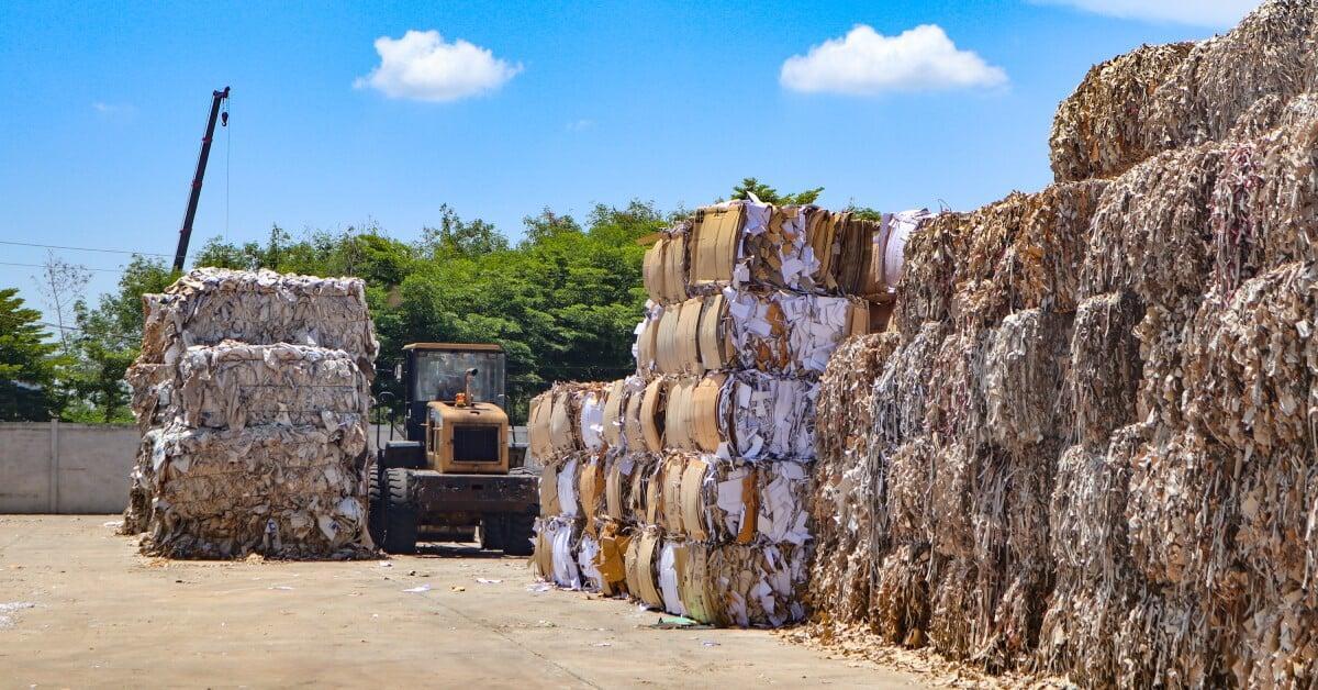 Lage bales of cardboard waste sit in tall stacks on an outdoor paved lot on a sunny day. A tractor sits nearby.