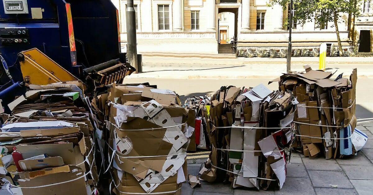 Bales of flattened cardboard boxes sit outside on a sidewalk near a large vertical cardboard baling machine.