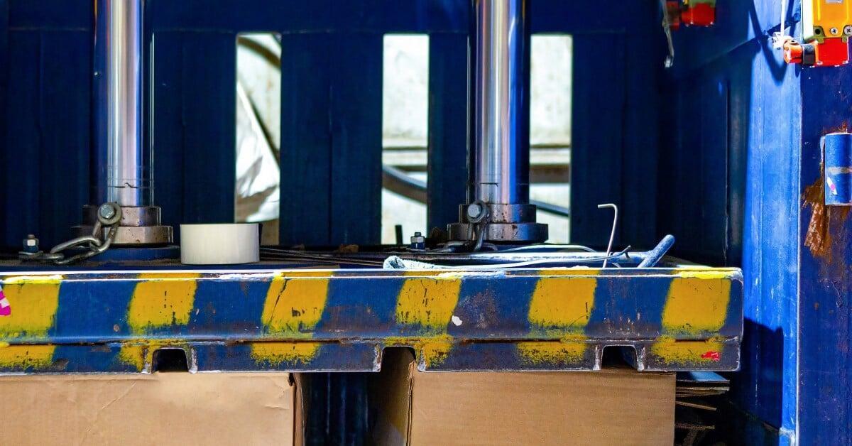  A view of the hydraulic components of a cardboard baling machine. Two boxes sit below the compression plate.