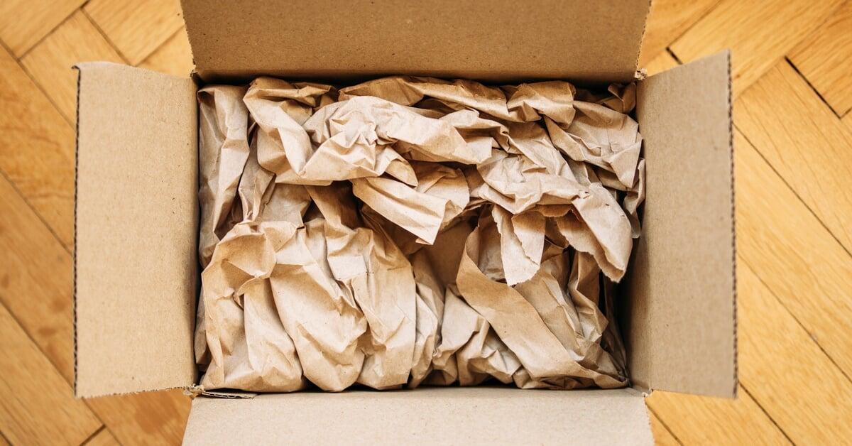 An open cardboard box sits on an oak hardwood floor. The box is stuffed with crumpled kraft paper void fill.