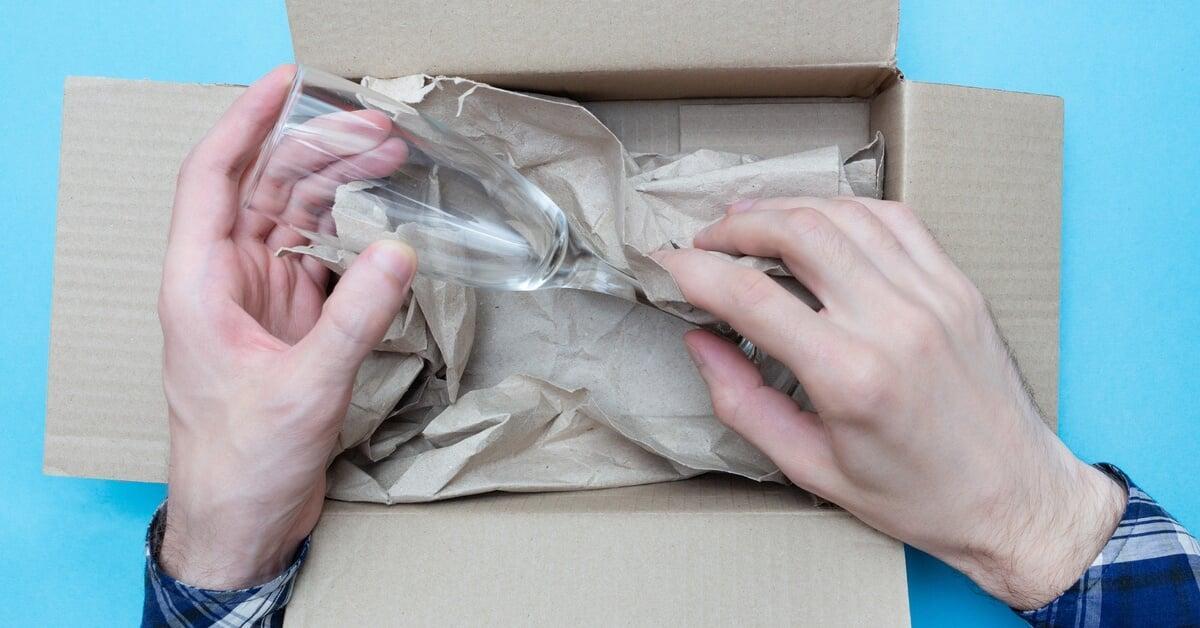 A person carefully places a clear wineglass into a small cardboard box that's filled with kraft paper.