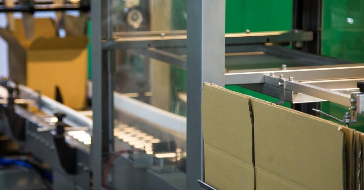 Flat cardboard boxes sit on one end of a case erector machine. The machine forms a box at the end of the line.