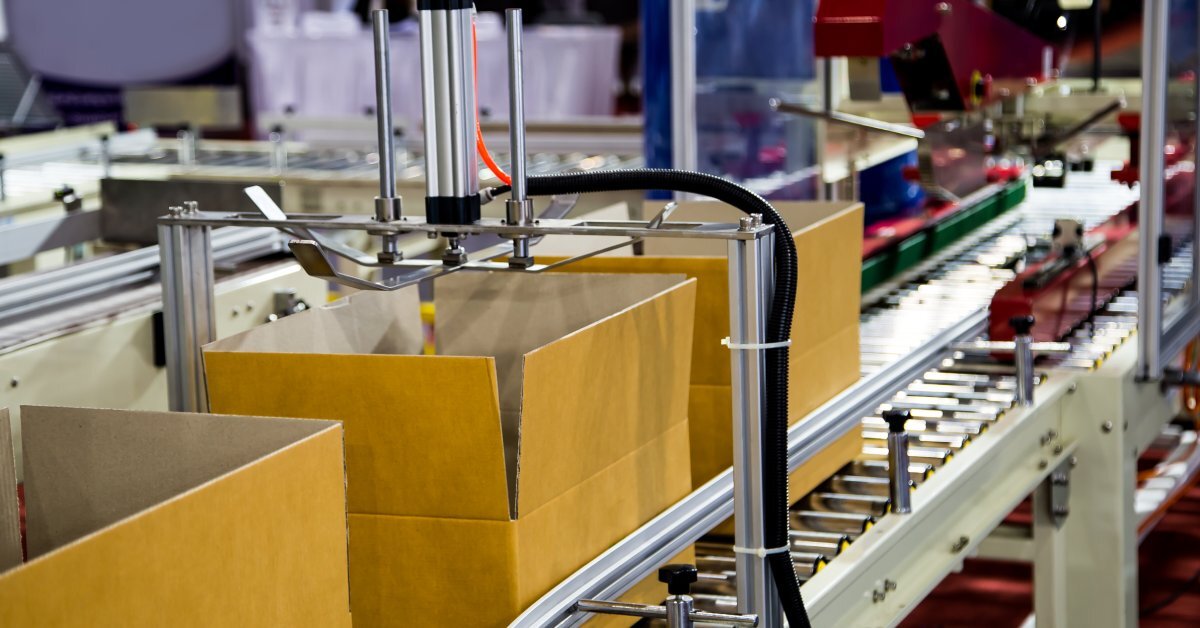 Three cardboard boxes with open top flaps move along the metal conveyor belt on a case former machine.
