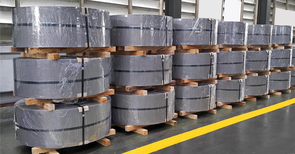 Large rolls of silver sheet metal are strapped to wooden pallets. The palletized rolls are stacked on top of each other.