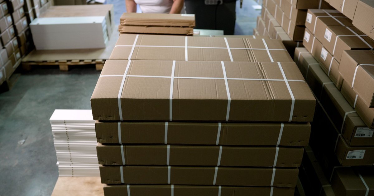 A stack of slim cardboard boxes are individually wrapped with plastic straps. The boxes sit on a wooden pallet.