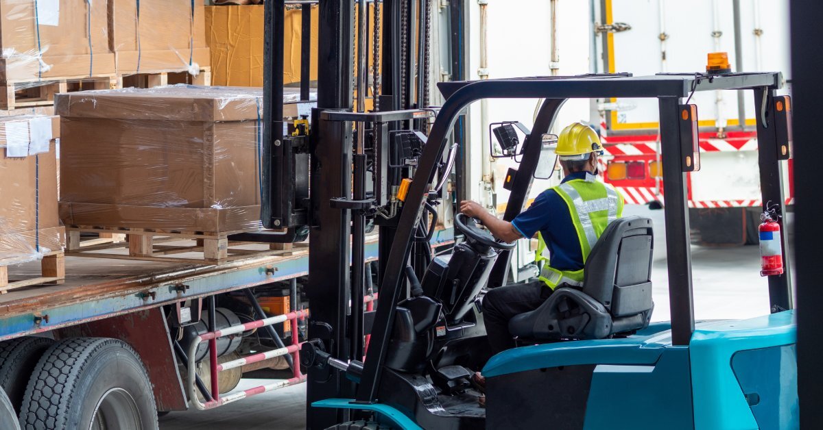 A warehouse worker uses a forklift to move a wrapped pallet of cardboard boxes off of a flat-bed trailer.