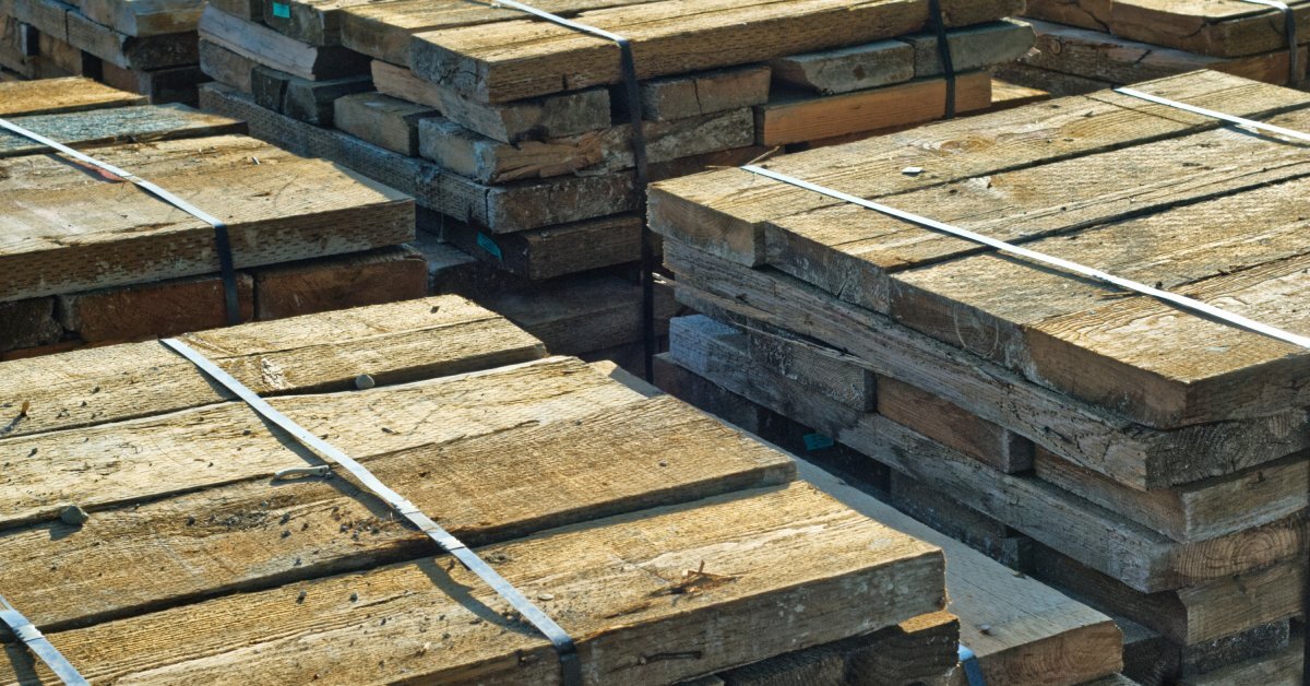 Large bundles of wide wooden planks are secured with metal strapping. They sit outside in the sunshine.