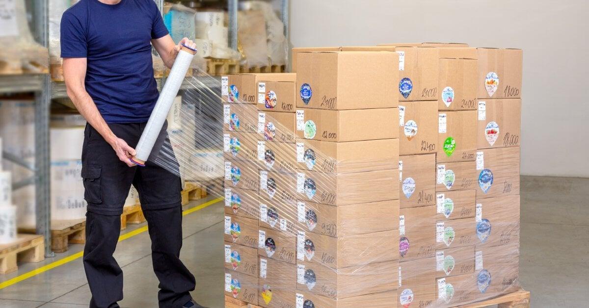 A man holds a roll of clear plastic film as he manually wraps a pallet stacked high with cardboard boxes.