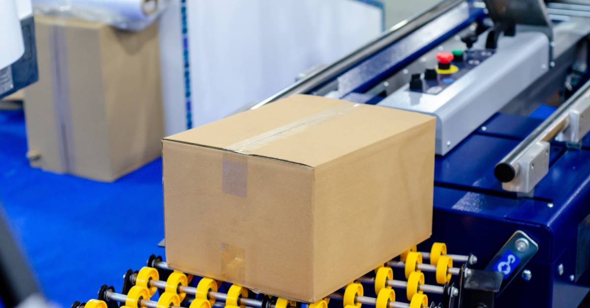 A sealed carton on a conveyor belt with yellow skate wheels. A box and packaging supplies sit in the background.