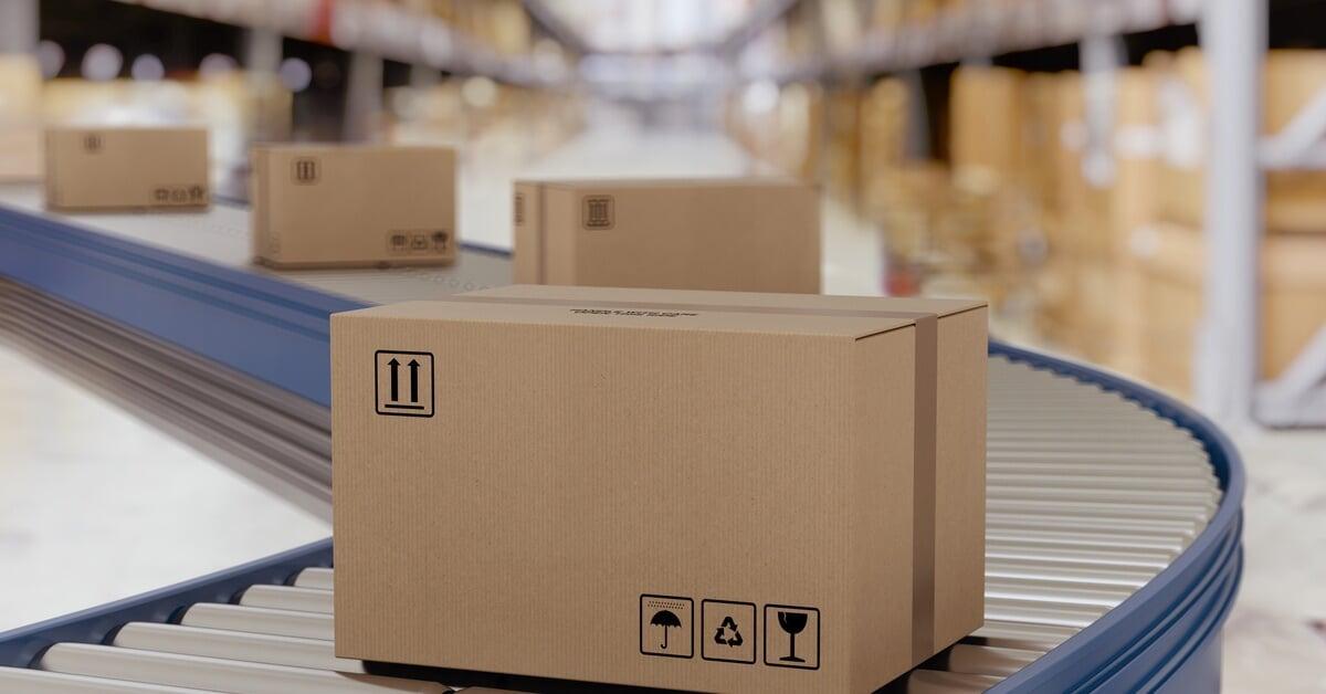 Four brown, cardboard boxes spaced apart sit on a conveyor belt; there are shelves of boxes blurred in the background.