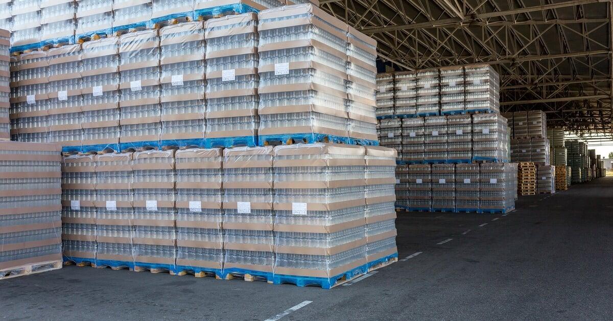 Several stacks of shrink-wrapped items sitting on blue pallet racks, all stacked on top of one another in a warehouse.