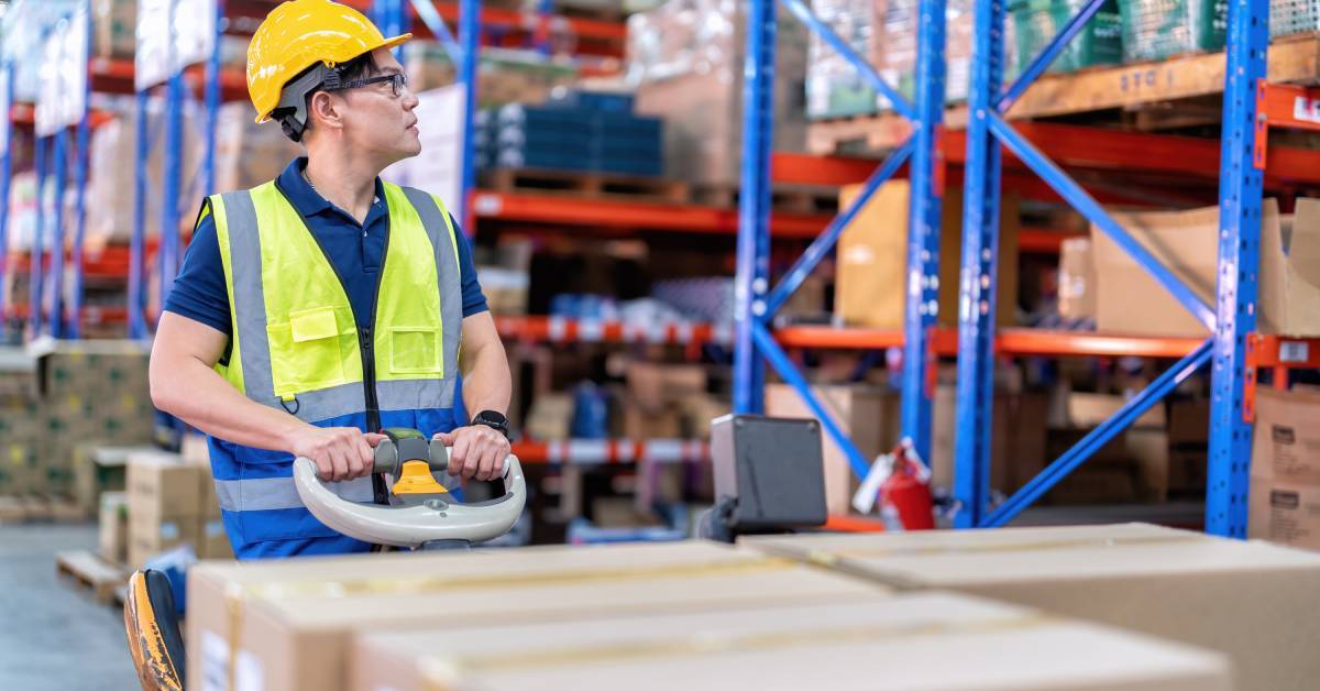 An employee operating a loaded pallet jack and looking up at shelves in a warehouse. Several shelves of goods are behind him.