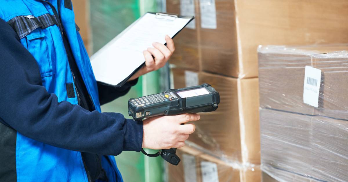 A worker scanning a barcode on a stretch-wrapped pallet of cardboard cases. They are holding a clipboard in their other hand.