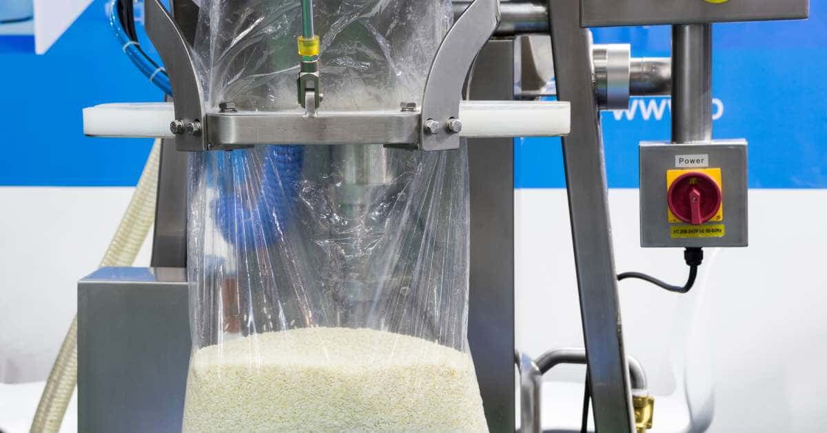 A machine in front of a blue and white wall holding open a thin plastic bag partially filled with uncooked rice.