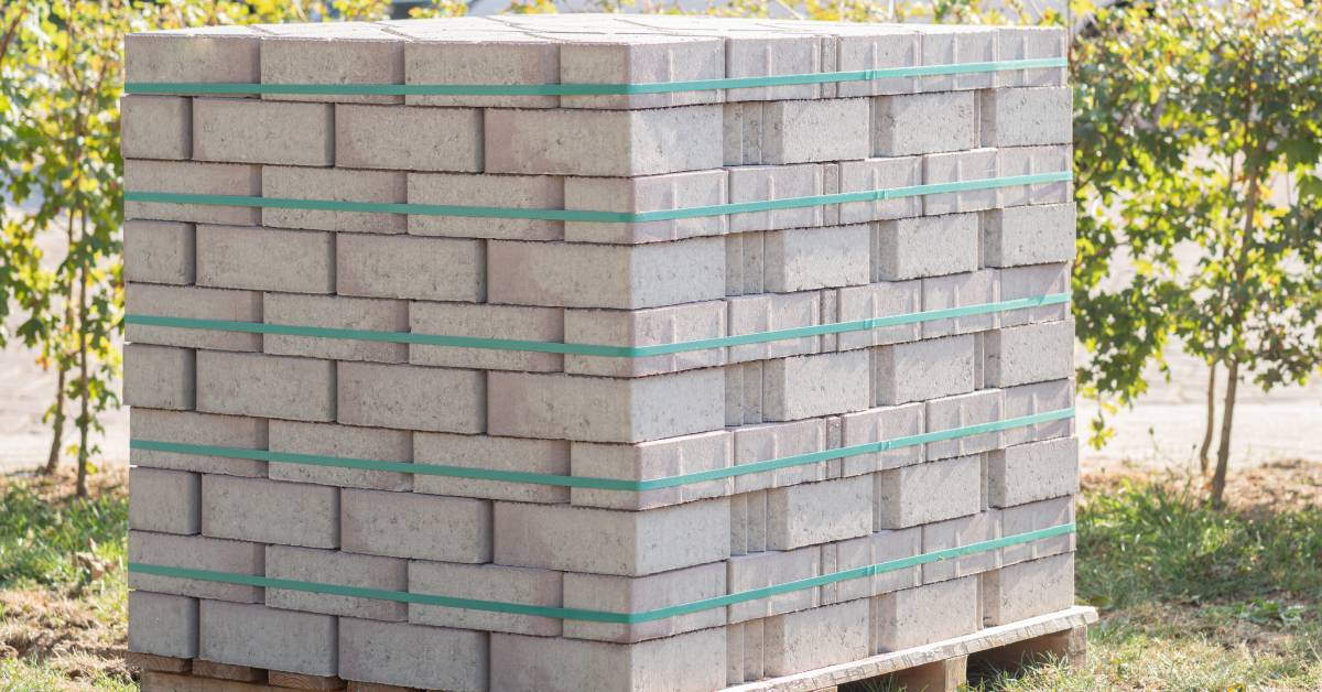 A pallet of concrete bricks stored outside on the grass. Five green straps wrapped around the bricks are evenly spaced apart.