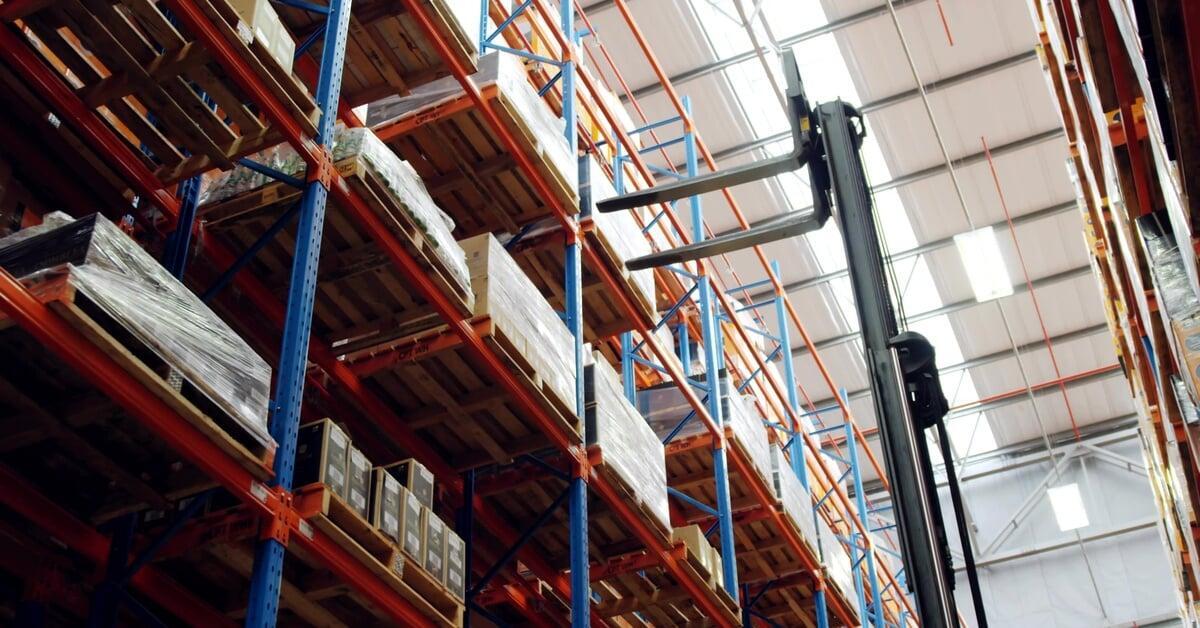 A high-bay warehouse with tall pallet racks and a forklift mast lifting pallets among stacked boxes.