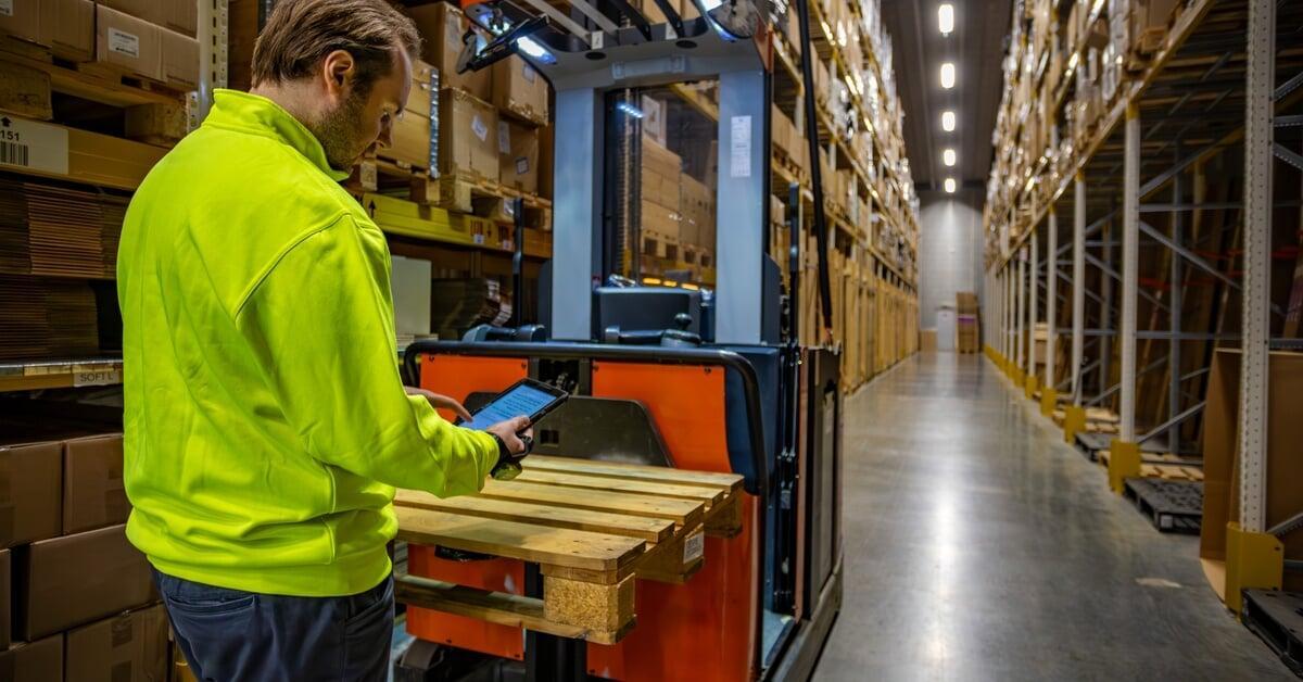 A warehouse worker in a safety jacket using a tablet to operate a pallet stacker in a storage aisle.